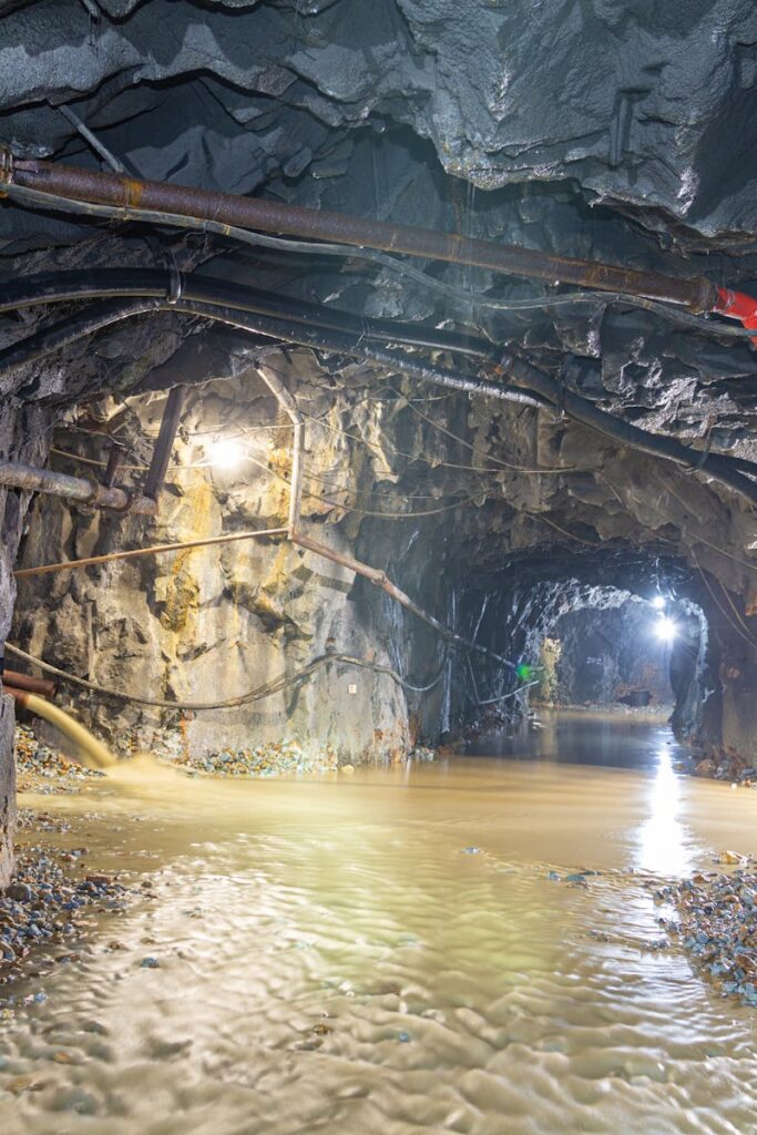 Dimly lit underground cave with flowing water and exposed pipes, showcasing rugged geology.