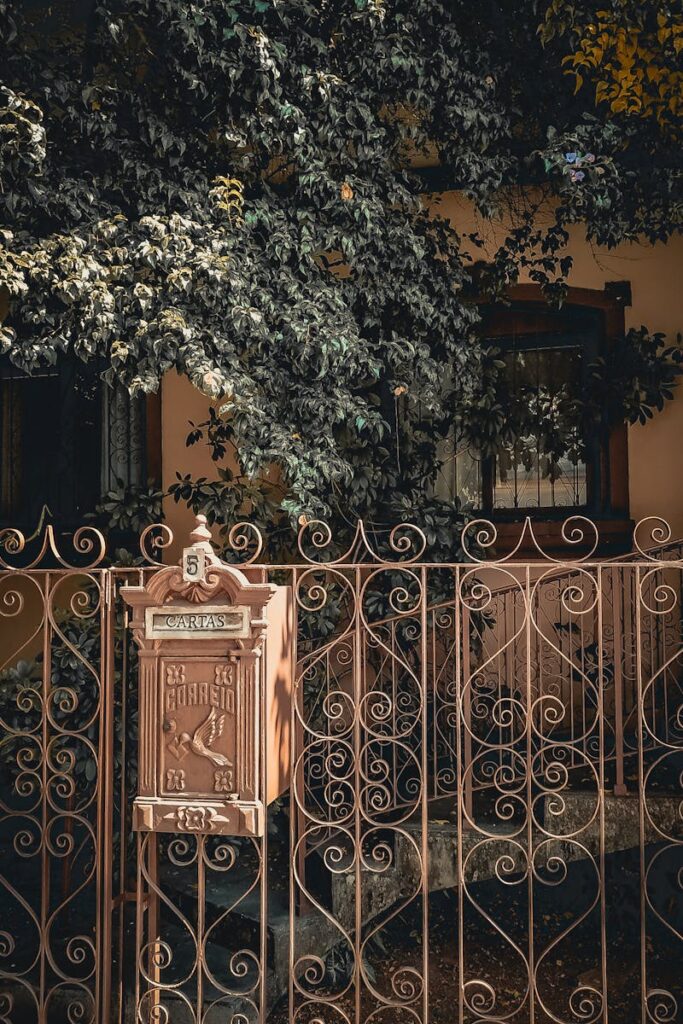 Ornate iron gate with mailbox, vintage design surrounded by lush foliage.