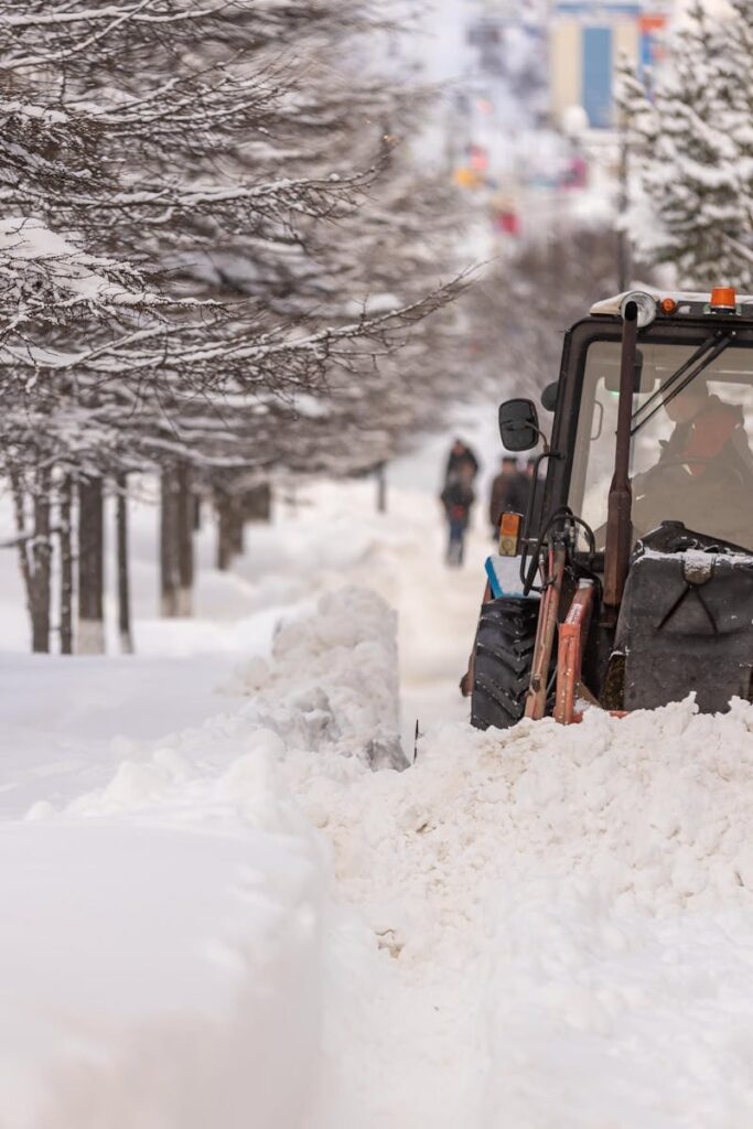 A snowplow truck clearing a snowy street in a winter urban setting with snow-covered trees.