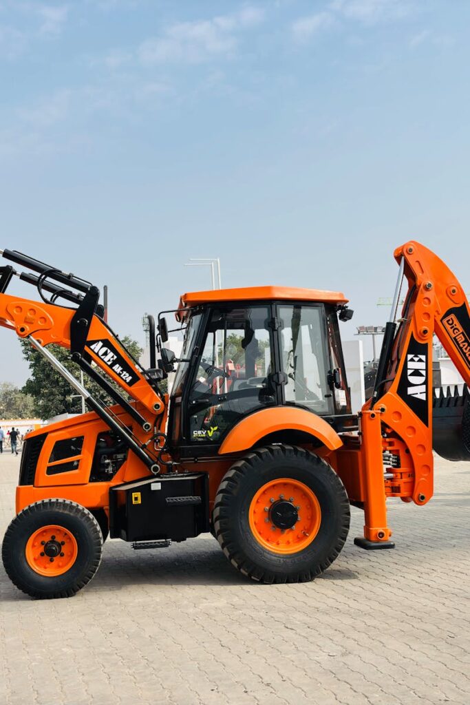 Bright orange ACE AX 124 backhoe loader on display at an outdoor exhibition in India.